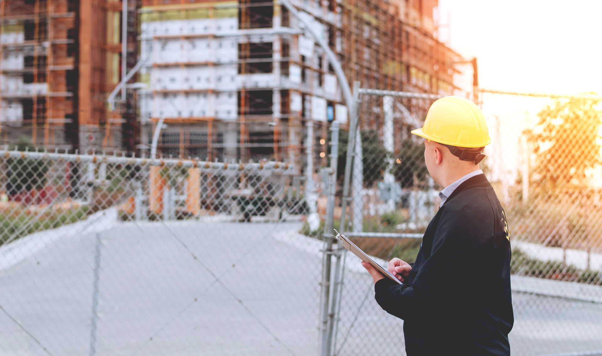 A man in a yellow hard hat, with a clipboard, by a barbed wire fence, gazing at a construction site.